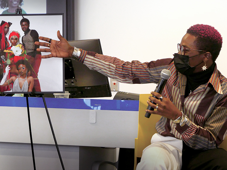 A masked person speaking into a microphone in front of a framed photograph